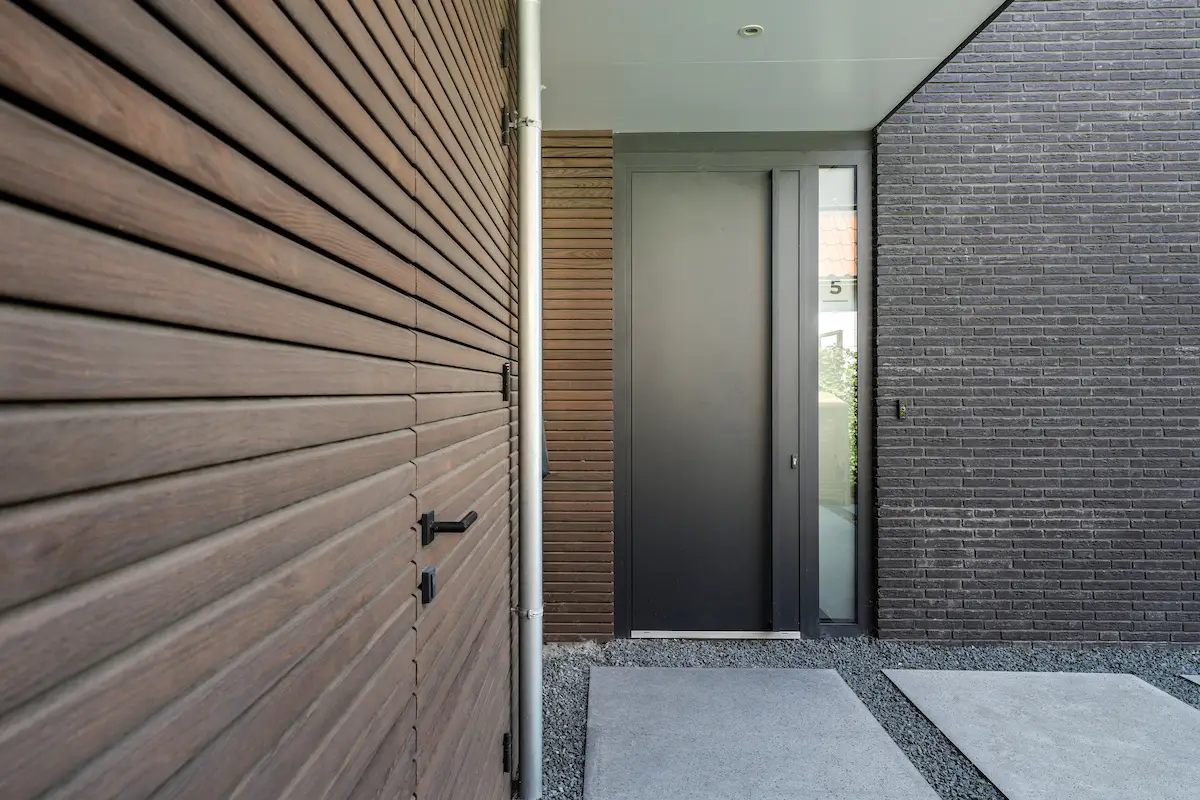 Beschutte entree van een moderne woning in Hendrik-Ido-Ambacht met houten gevelbekleding, donkere baksteen en een hoge voordeur met zijlicht.