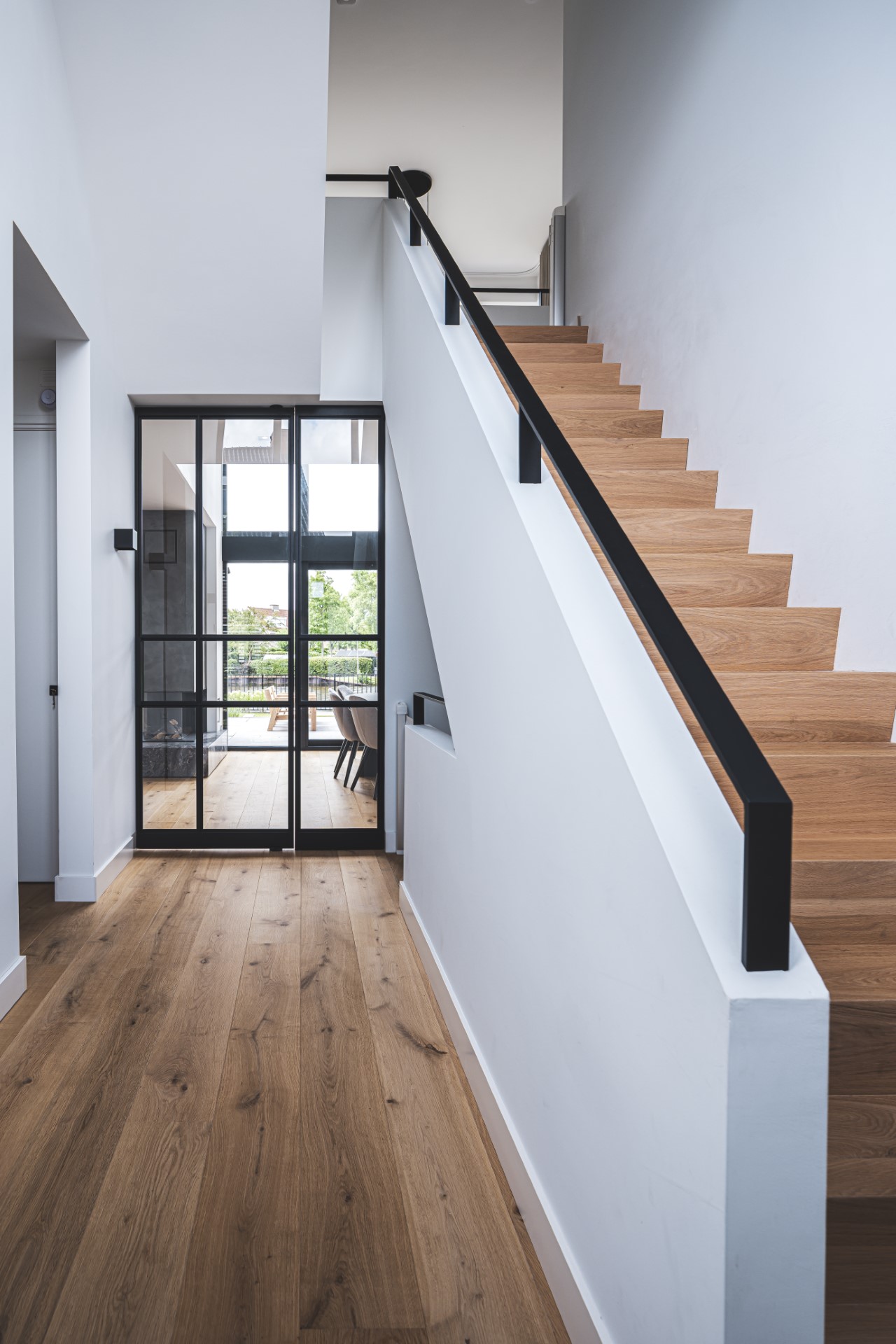 Lichte entree met houten trap, stalen taatsdeur en eiken vloer in duurzaam ontworpen moderne woning.