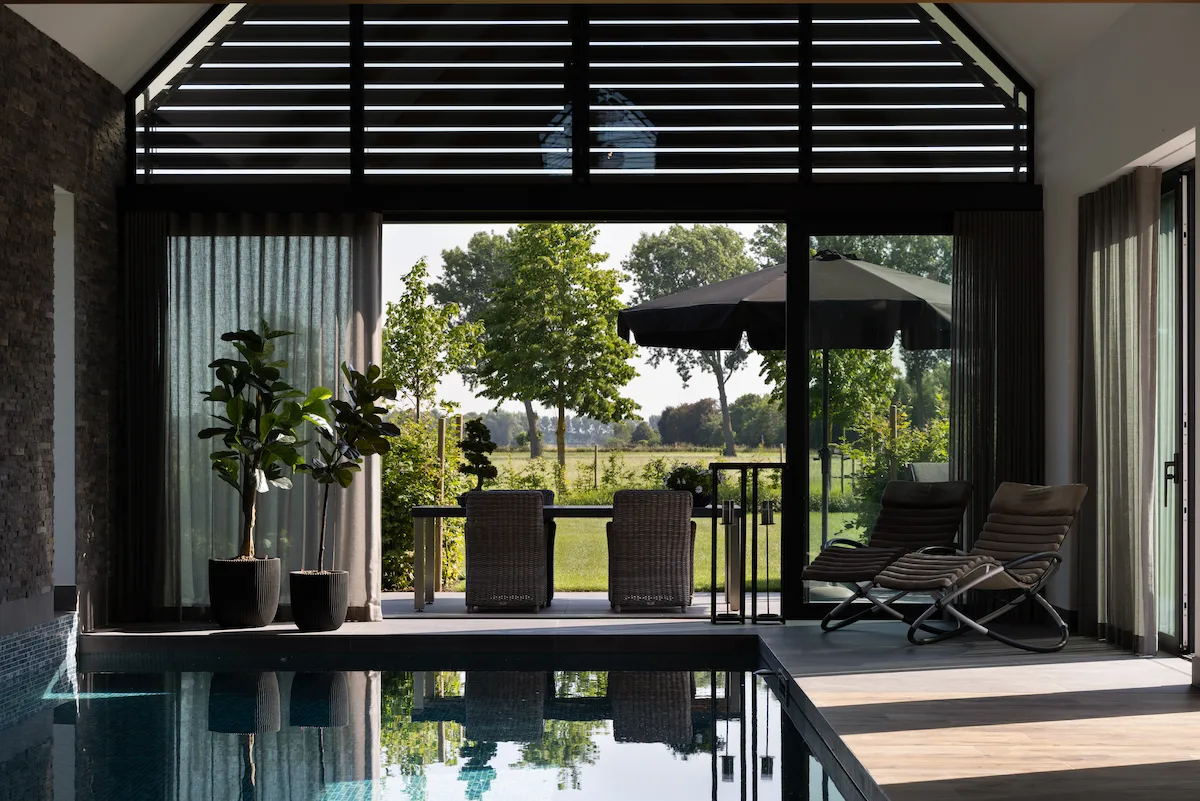 Binnenzwembad met grote glaspartijen en zwarte lamellen, met vrij zicht op de groene tuin en het omliggende landschap.