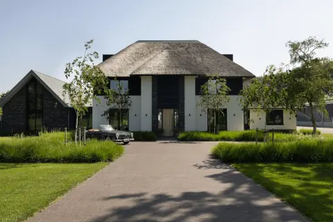 Moderne villa met rieten kap, witte gevels en zwarte houten accenten, stijlvol ingepast in een groene tuin met lange oprijlaan.