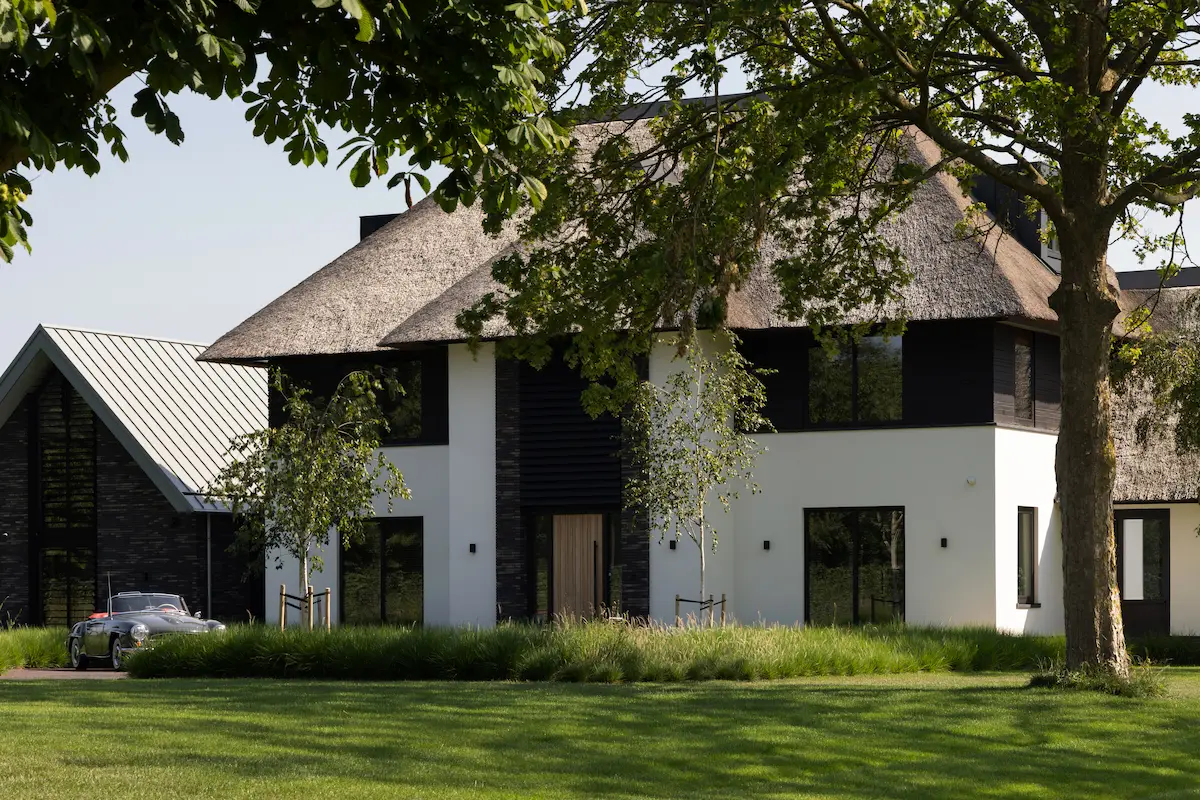 Moderne villa met rieten kap, witte gevel en zwarte houten accenten, natuurlijk ingepast in een groene tuin met volwassen bomen.