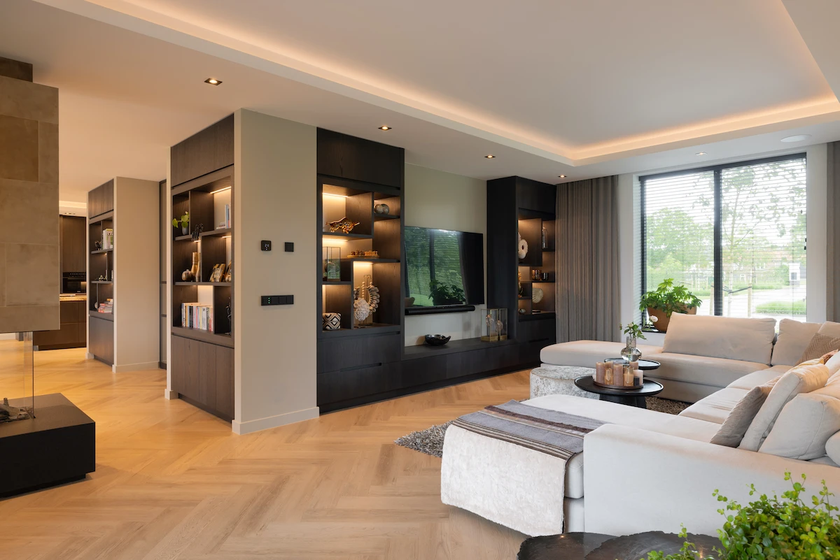Lichte woonkamer met maatwerk wandkast, visgraatvloer en grote glaspartijen, uitgevoerd als warm en minimalistisch interieur.