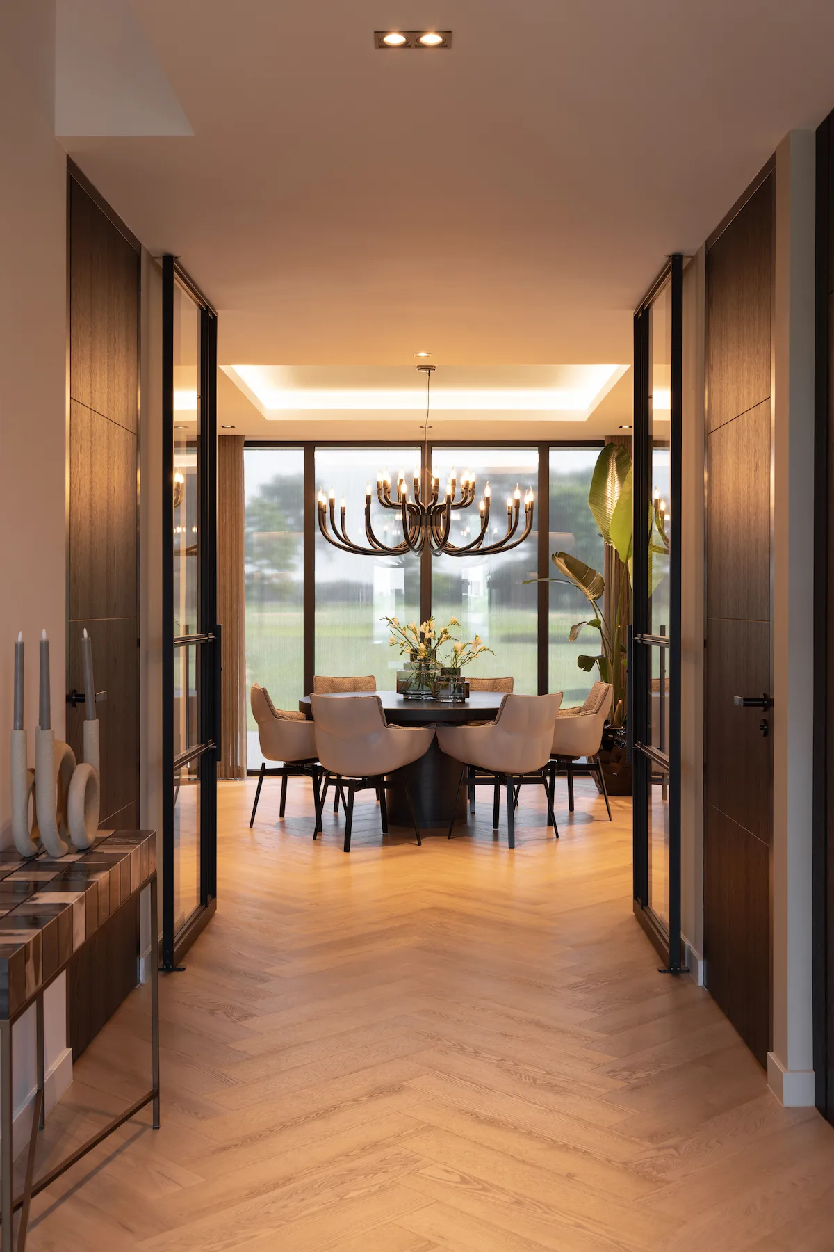 Luxe eetkamer met visgraatvloer, maatwerk deuren en grote glaspartijen, met warme verlichting en vrij zicht op het groene landschap.