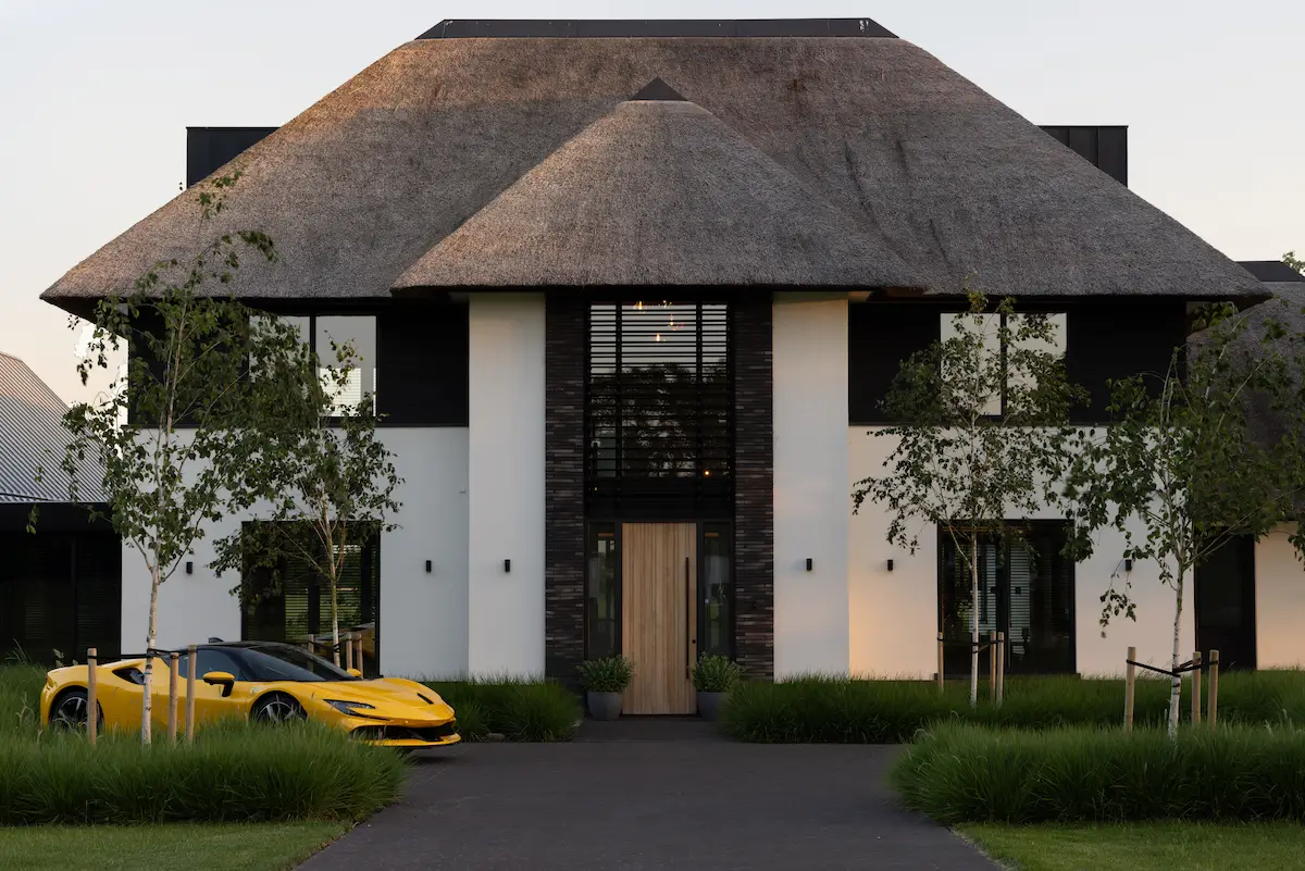 Moderne villa met rieten kap, symmetrische witte gevel en zwarte accenten, met hoge entree en groene landschappelijke tuin.