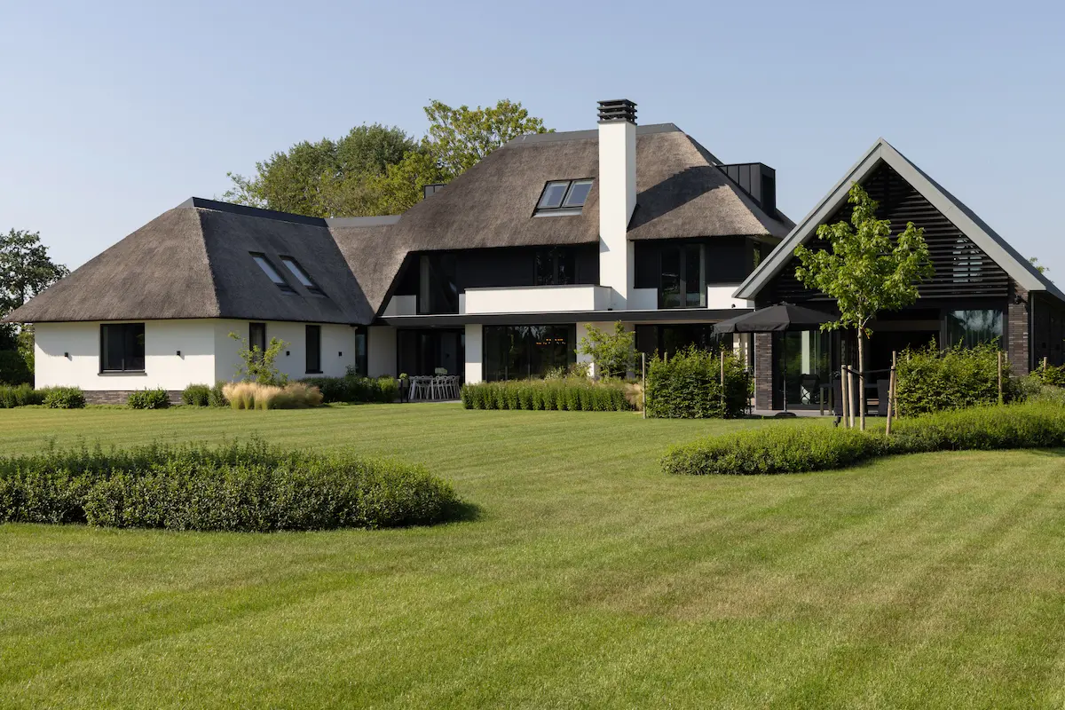 Moderne villa met rieten kap, grote glaspartijen en zwart houten bijgebouw, omringd door een ruime tuin met strakke groene borders.