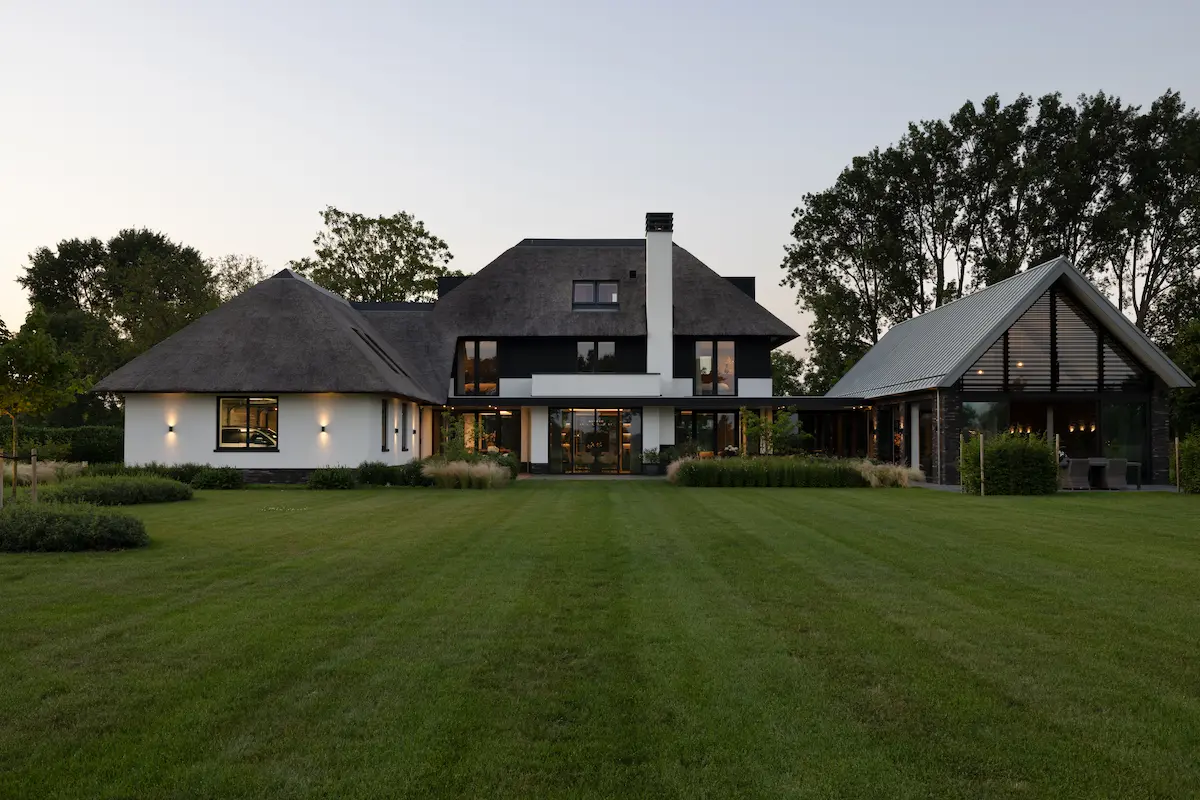 Moderne villa met rieten kap, grote glaspartijen en bijgebouw in een groene tuin, met strakke architectuur en sterke zichtlijnen.