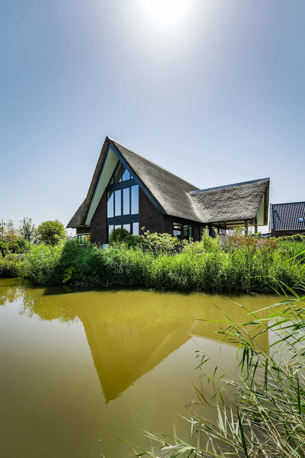 Moderne villa aan het water met rieten kap, hoge glasgevel en weerspiegeling in de vijver, ontworpen door architect Molenlanden.